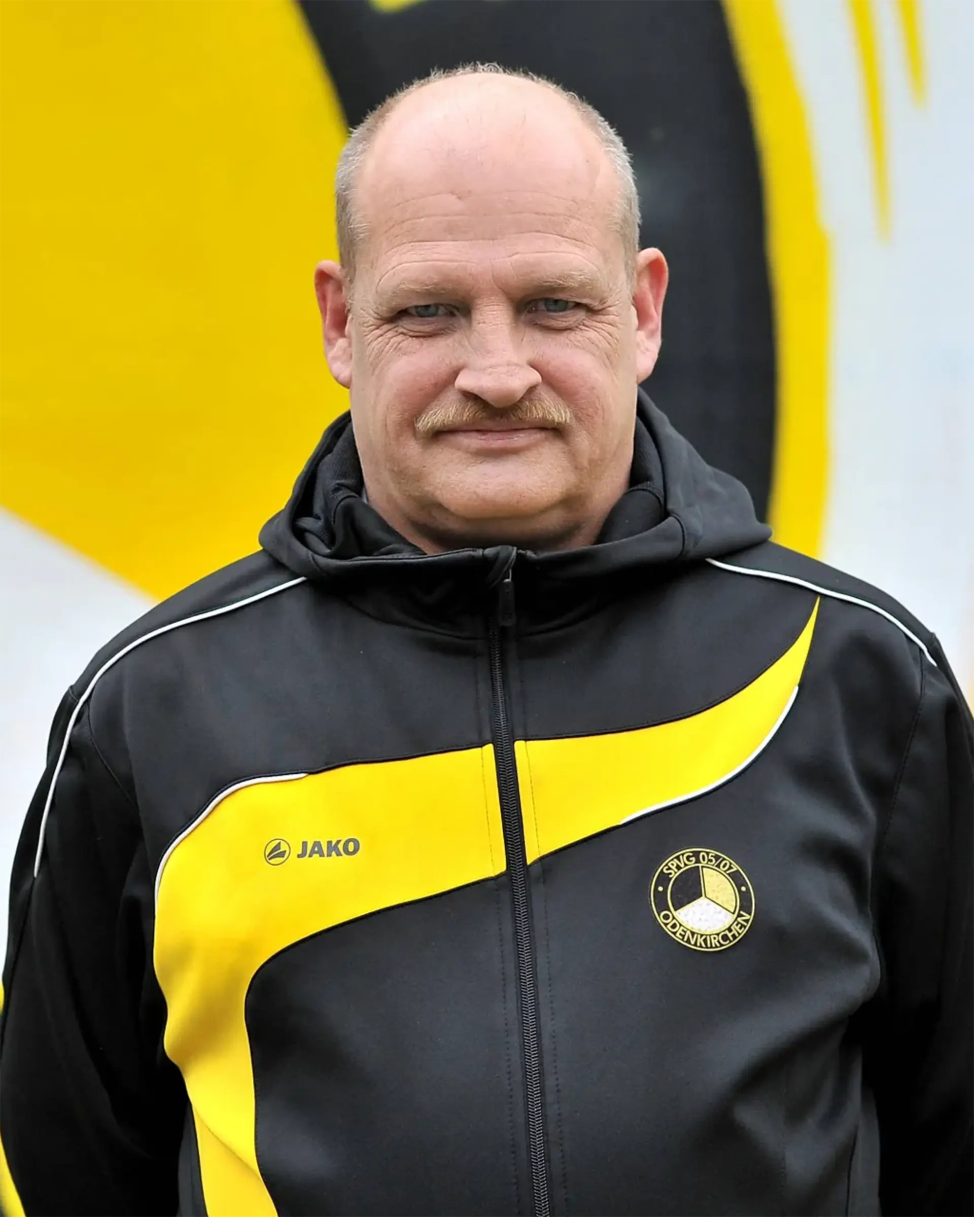 Ein Mann mittleren Alters mit Schnurrbart und schwarz-gelber Sportjacke steht vor einer passenden gelb-schwarzen Wand - und repräsentiert stolz den Fußball Alte Herren.