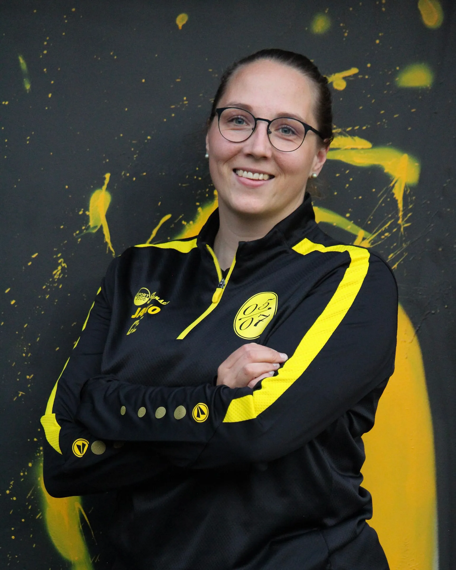 Eine Person mit Brille und braunen, nach hinten gezogenen Haaren lächelt mit verschränkten Armen, trägt eine schwarz-gelbe Fußball-Damen-Sportjacke mit einem gelben kreisförmigen Logo und steht vor einem schwarz-gelben abstrakten Hintergrund.