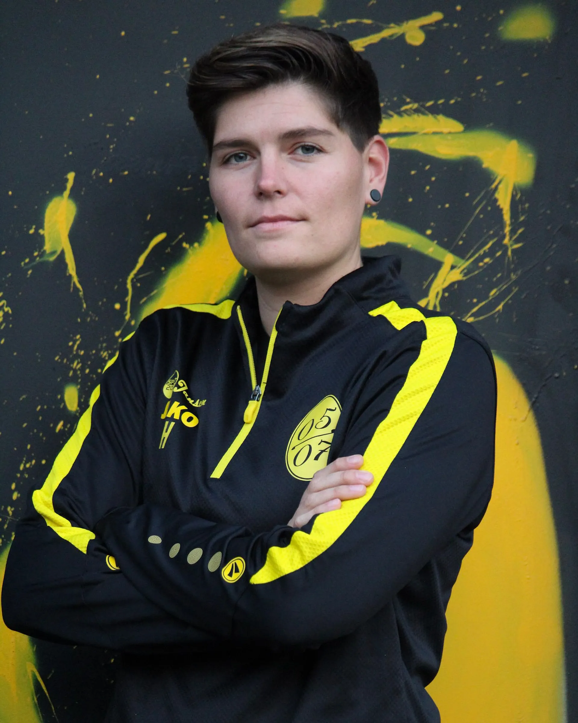 Eine Person mit kurzen braunen Haaren, die eine schwarz-gelbe Sportjacke mit 09-Logo trägt, steht selbstbewusst mit verschränkten Armen vor einer schwarz-gelben Graffiti-Wand und verkörpert den Geist der Fußball Damen.
