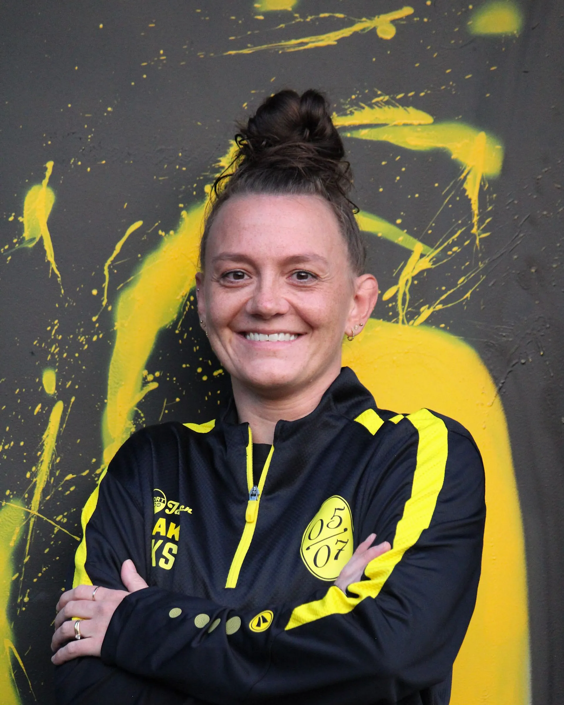 Lächelnde Person mit braunen Haaren in einem Haarknoten, die eine schwarz-gelbe Fußball Damen Sportjacke mit einem 05 07 Logo trägt, steht mit verschränkten Armen vor einem dunklen Hintergrund mit leuchtend gelben Farbspritzern.