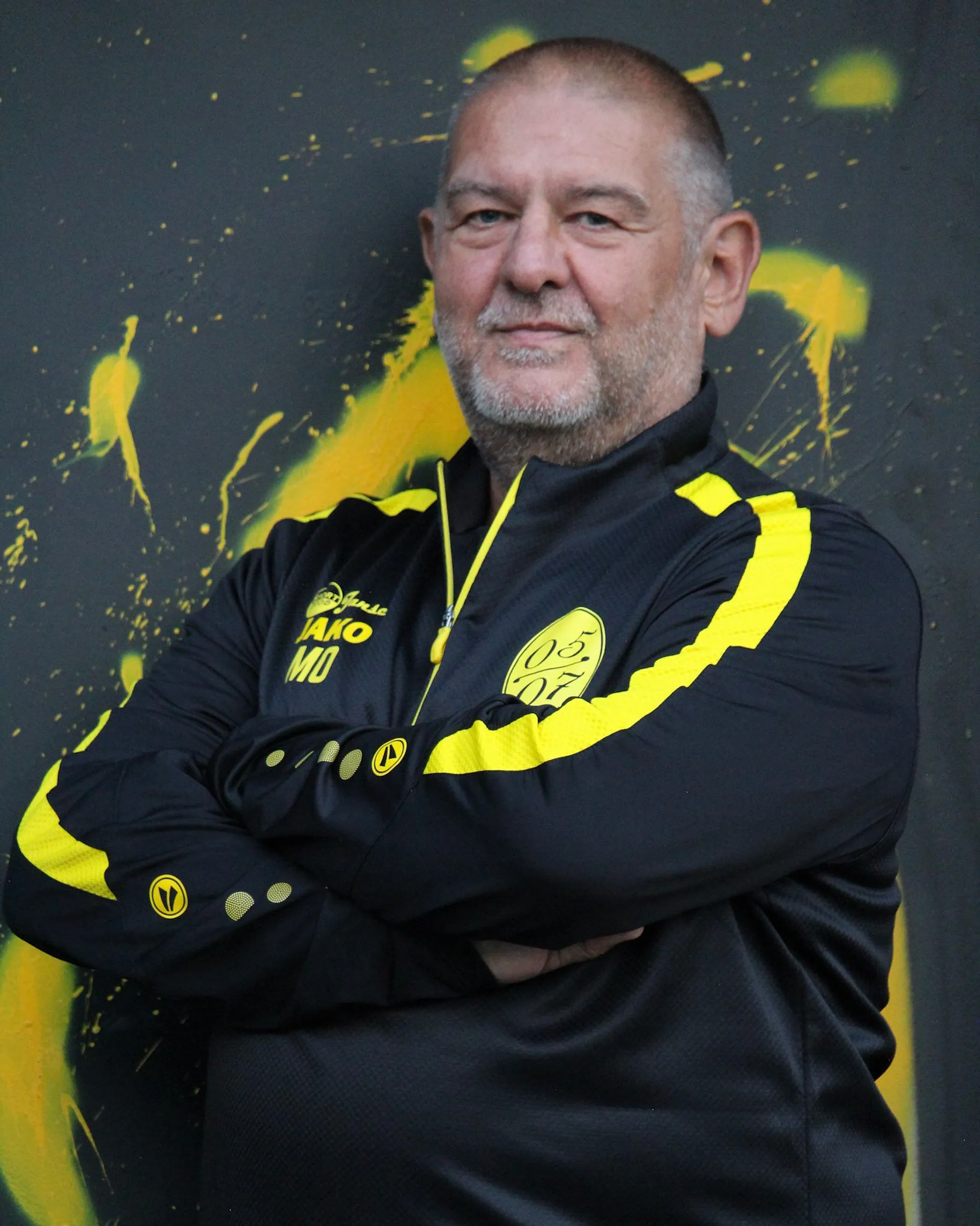 Ein Mann mit kurzen grauen Haaren und Bart steht mit verschränkten Armen in einer schwarz-gelben Fußball-Damen-Sportjacke mit Logos vor einem schwarzen, mit gelber Farbe bespritzten Hintergrund.