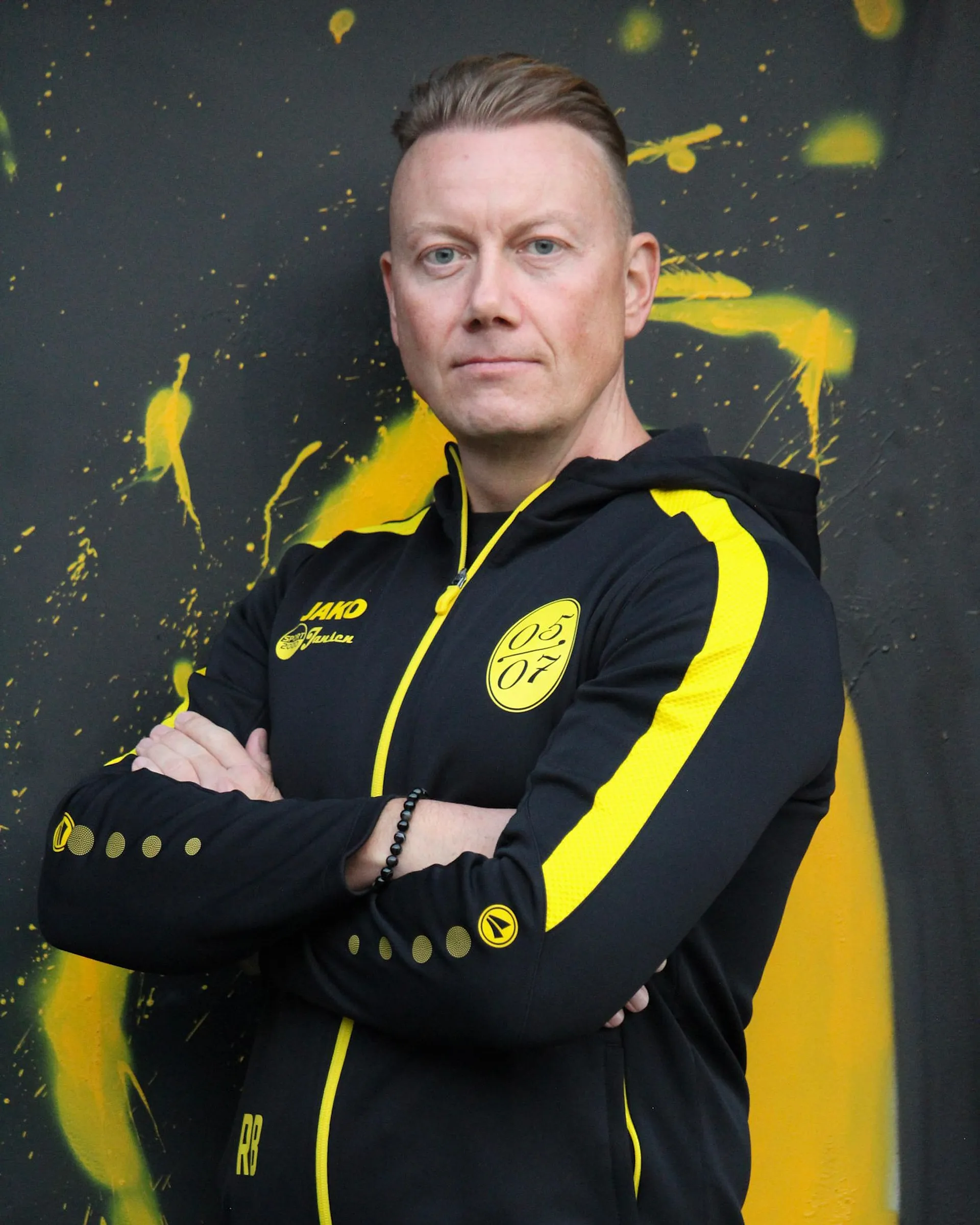 Ein Mann mit kurzen blonden Haaren steht mit verschränkten Armen und trägt eine schwarz-gelbe Sportjacke mit der Nummer 05 darauf. Er steht vor einem dunklen Hintergrund mit gelben Farbspritzern, die an die Energie von Fußball-Damenmannschaften erinnern.