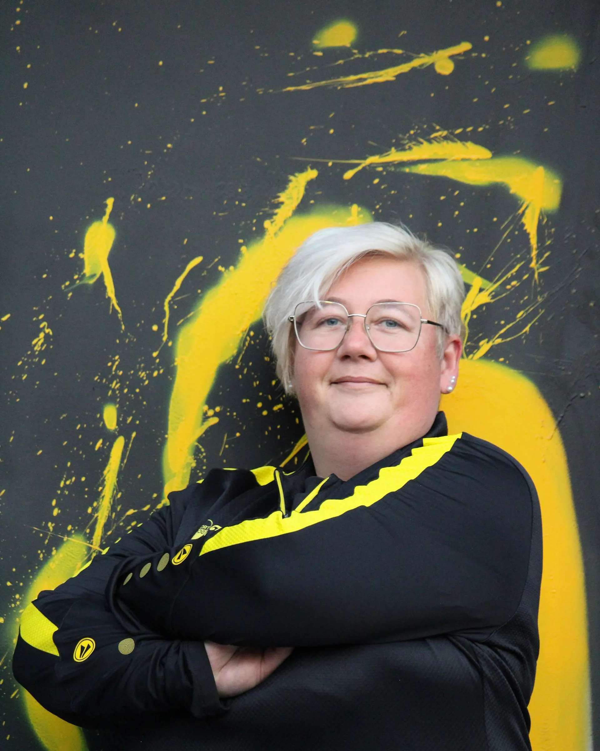 Eine Person mit kurzen weißen Haaren und Brille steht selbstbewusst mit verschränkten Armen in einer schwarz-gelben Fußball-Damen-Jacke vor einer grauen Wand mit kräftigen gelben Farbspritzern.