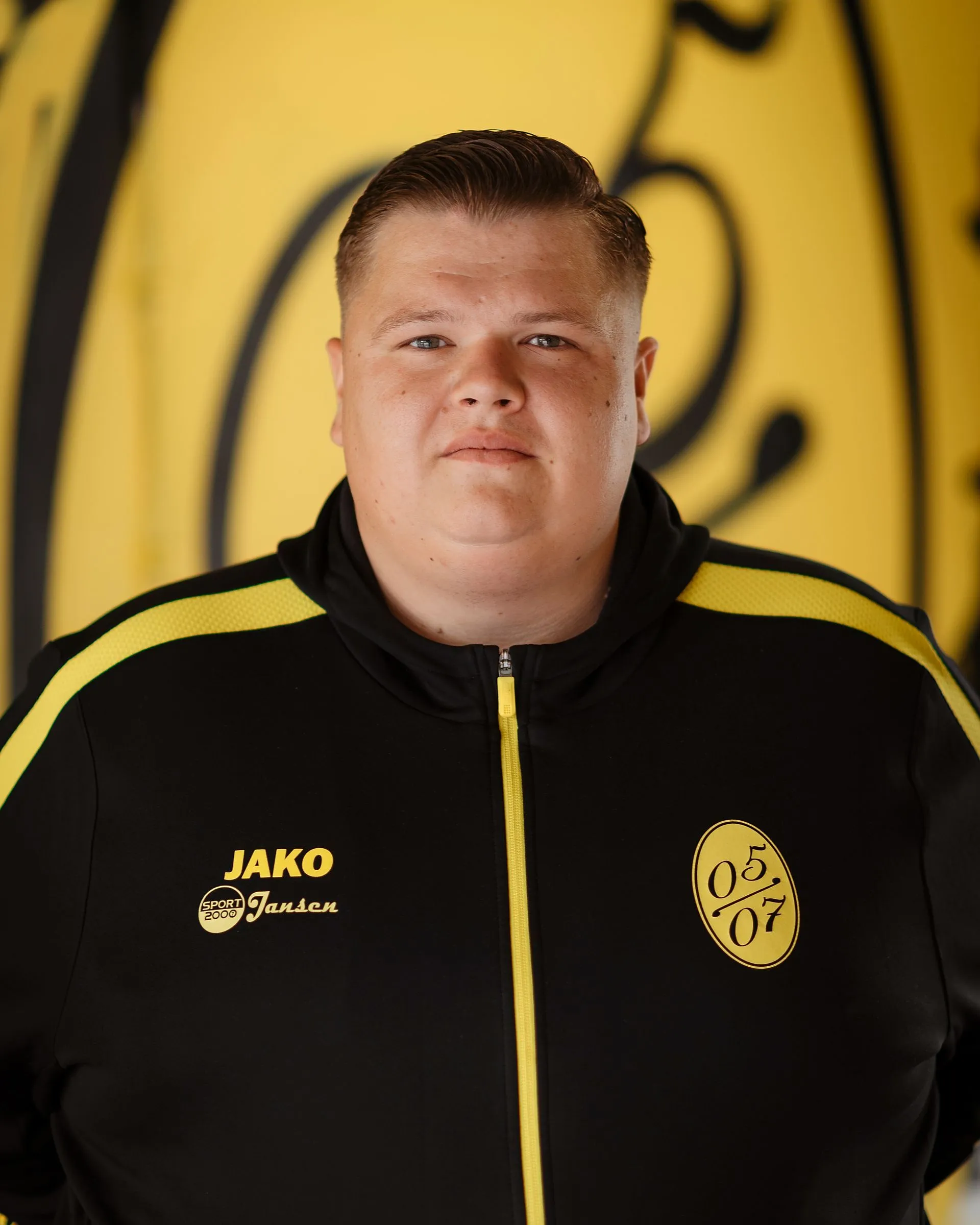 Ein Mann, der eine schwarz-gelbe Sportjacke mit den Logos von JAKO, Jansen und 05 07 trägt, steht als Vertreter der Fußball-Herren vor einem gelben Hintergrund mit schwarzen Motiven. Er blickt mit neutralem Blick direkt in die Kamera.