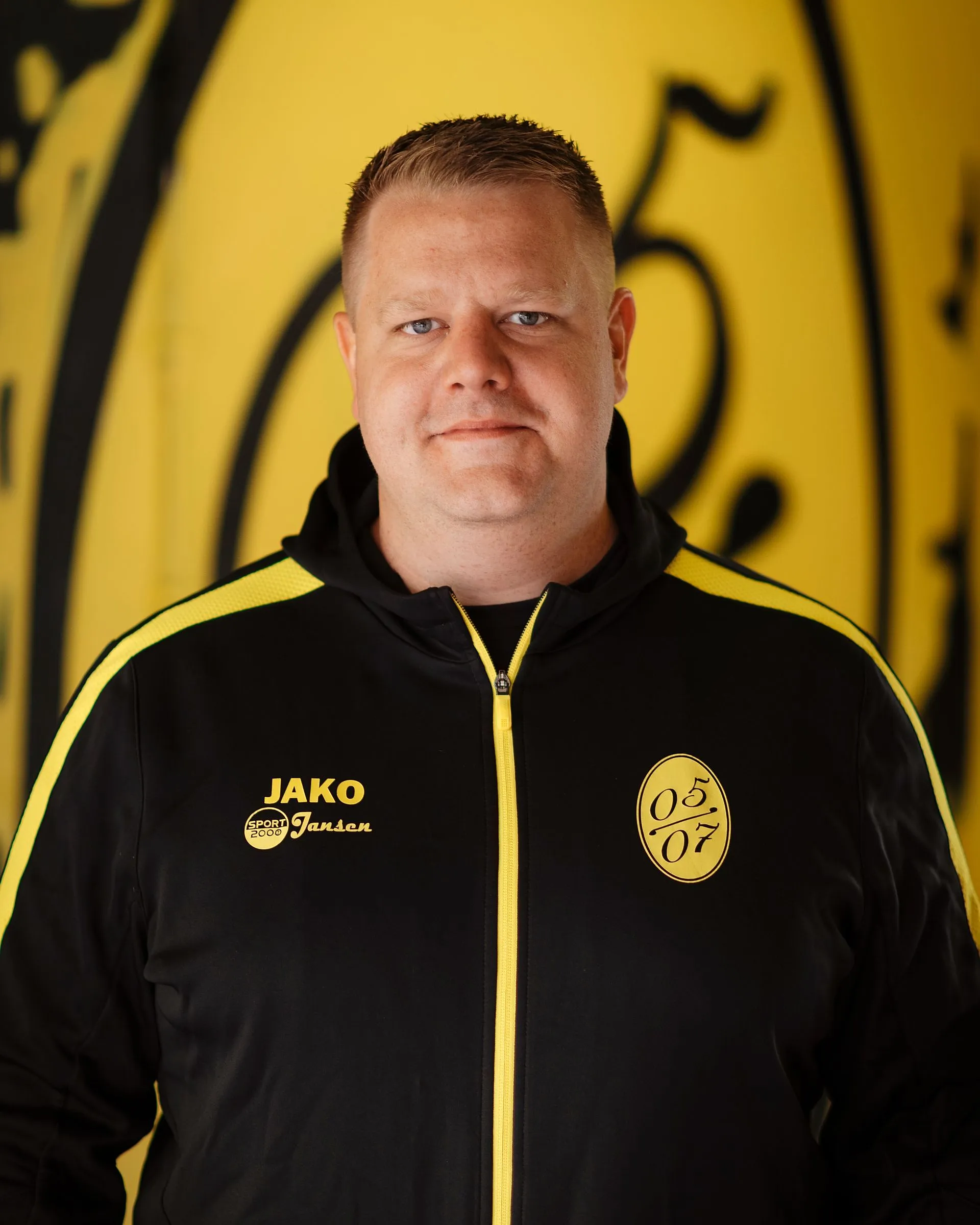 Ein Mann mit kurzen blonden Haaren, der eine schwarz-gelbe Fußball-Herrenjacke mit den Logos JAKO und 05 07 trägt, steht vor einem gelben Hintergrund mit einem großen schwarzen Muster.