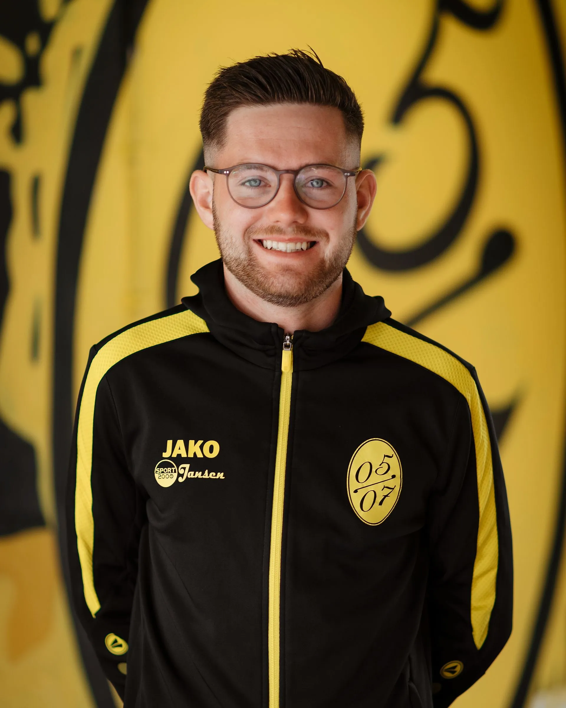 Ein lächelnder Mann mit kurzen braunen Haaren, Bart und Brille trägt eine schwarz-gelbe Fußball-Herren-Sportjacke mit JAKO- und 05/07-Logos und steht vor einem gelb-schwarzen abstrakten Hintergrund.