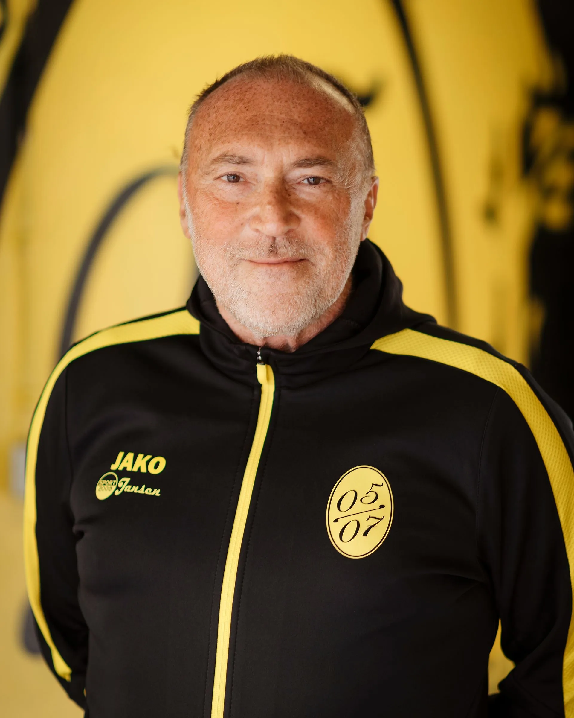 Ein Mann mit kurzen grauen Haaren und einem Bart lächelt in die Kamera. Er trägt eine schwarz-gelbe Fußball-Herren-Sportjacke mit Jako und 05 07-Logos und steht vor einem gelb-schwarzen Hintergrund.