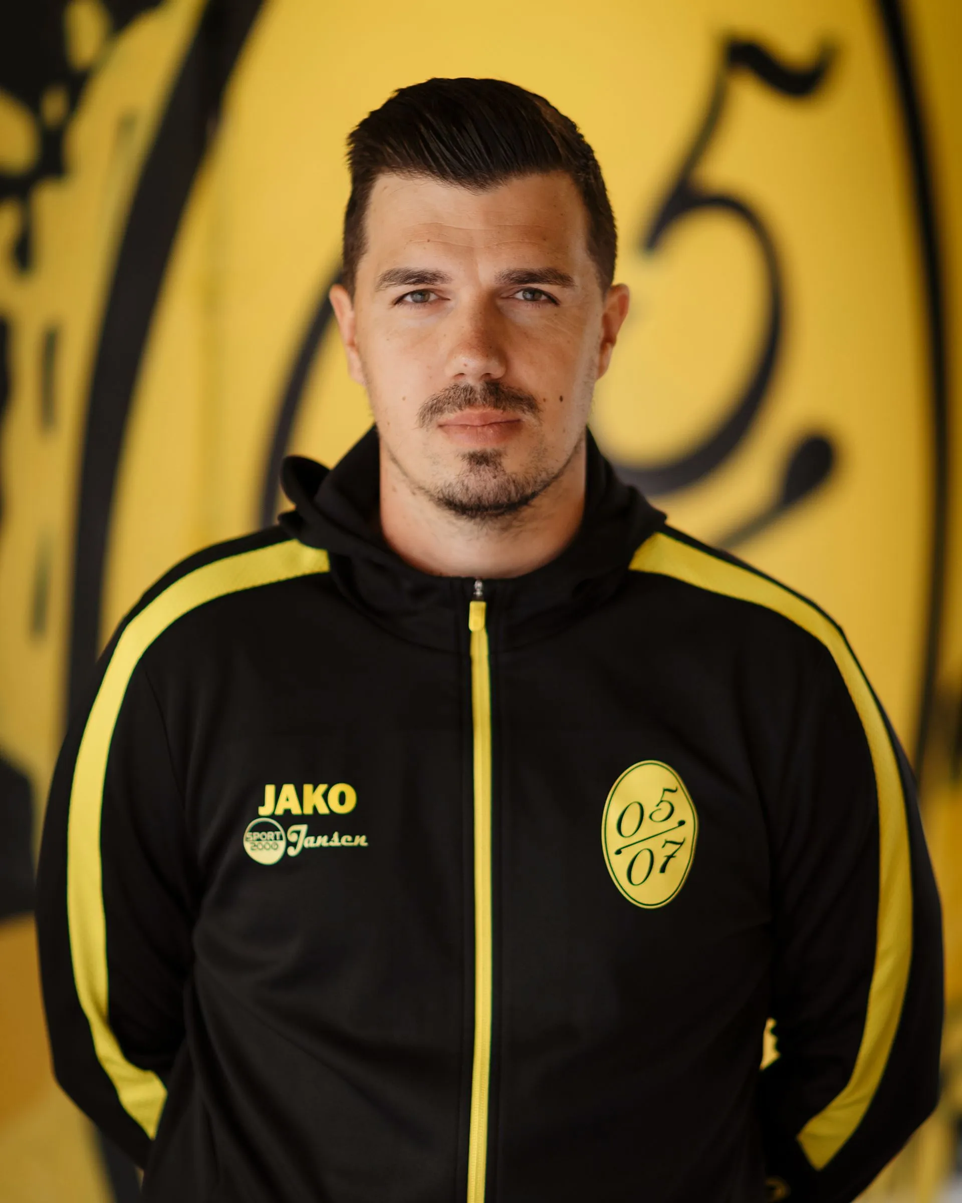 Ein Mann mit kurzen dunklen Haaren und gestutztem Bart, der eine schwarz-gelbe Fußball-Herren-Sportjacke mit einem 05/07-Emblem trägt, steht vor einer gelben Wand mit abstrakten schwarzen Motiven.