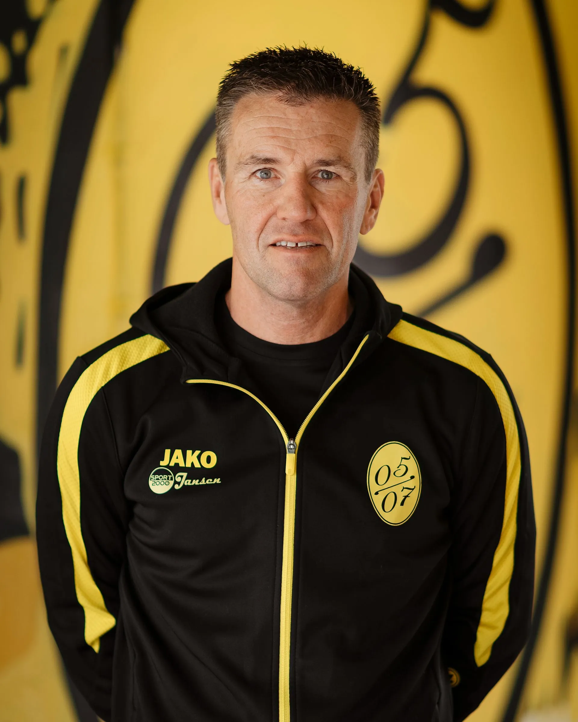 Ein Mann in einer schwarz-gelben Sportjacke mit einem JAKO-Logo und der Nummer 05 07, die für Fußball Herren steht, steht vor einem gelben Hintergrund mit schwarzen Motiven. Er hat kurzes braunes Haar und blickt in die Kamera.