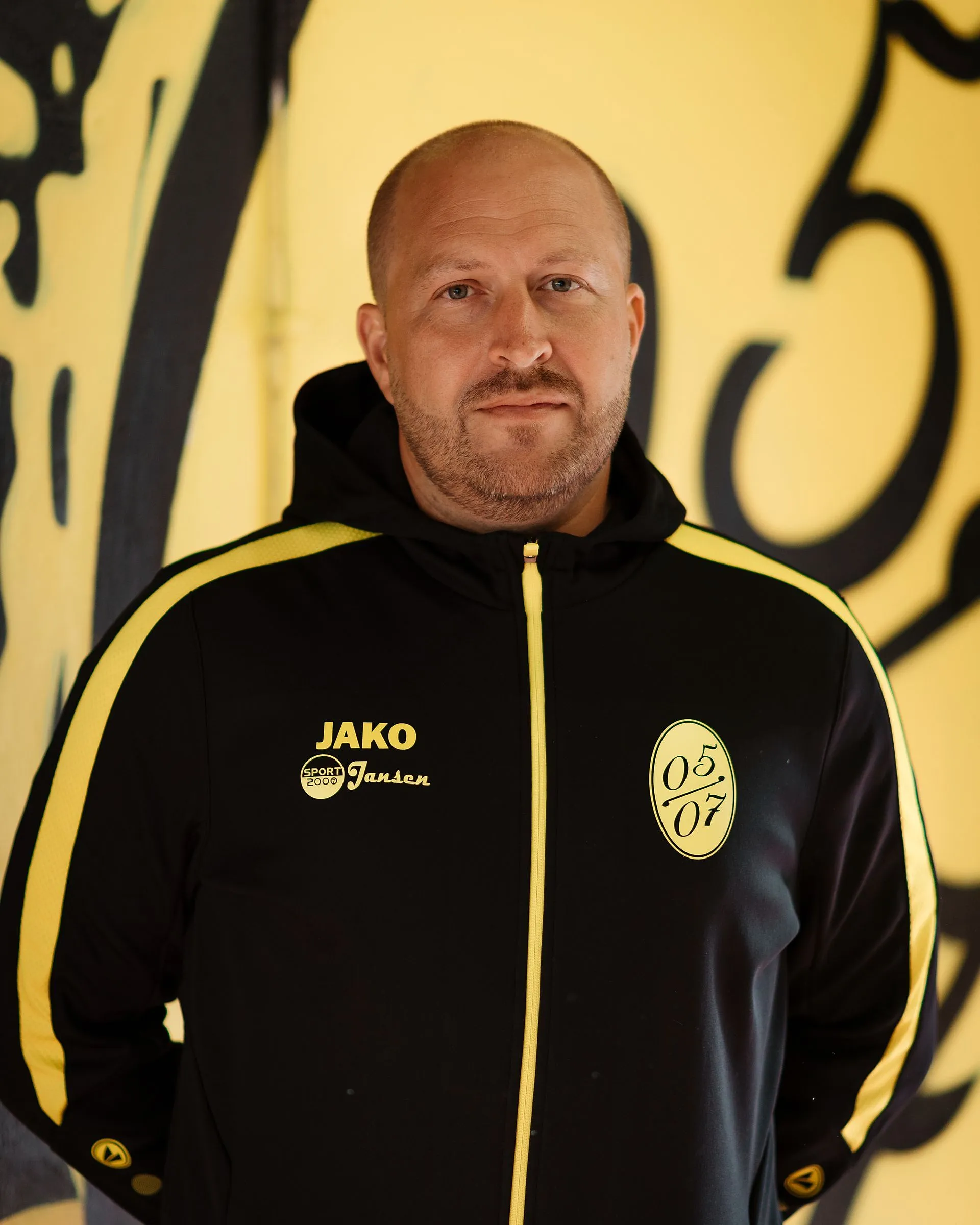 Ein Mann mit rasiertem Kopf und Bart trägt eine schwarz-gelbe Sportjacke mit JAKO-Logo, 05 07-Abzeichen und Fußball Herren 2-Emblem und steht vor einer gelben Wand mit schwarzen Motiven.