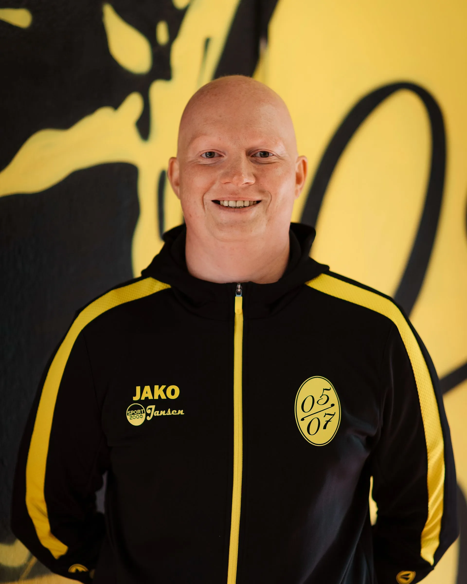 Ein lächelnder Mann mit Glatze in einer schwarz-gelben Sportjacke steht vor einer gelb-schwarzen Wandmalerei. Die Jacke zeigt den Text JAKO, Jansen und ein kreisförmiges Logo mit 05 07, das für Fußball Herren 2 steht.