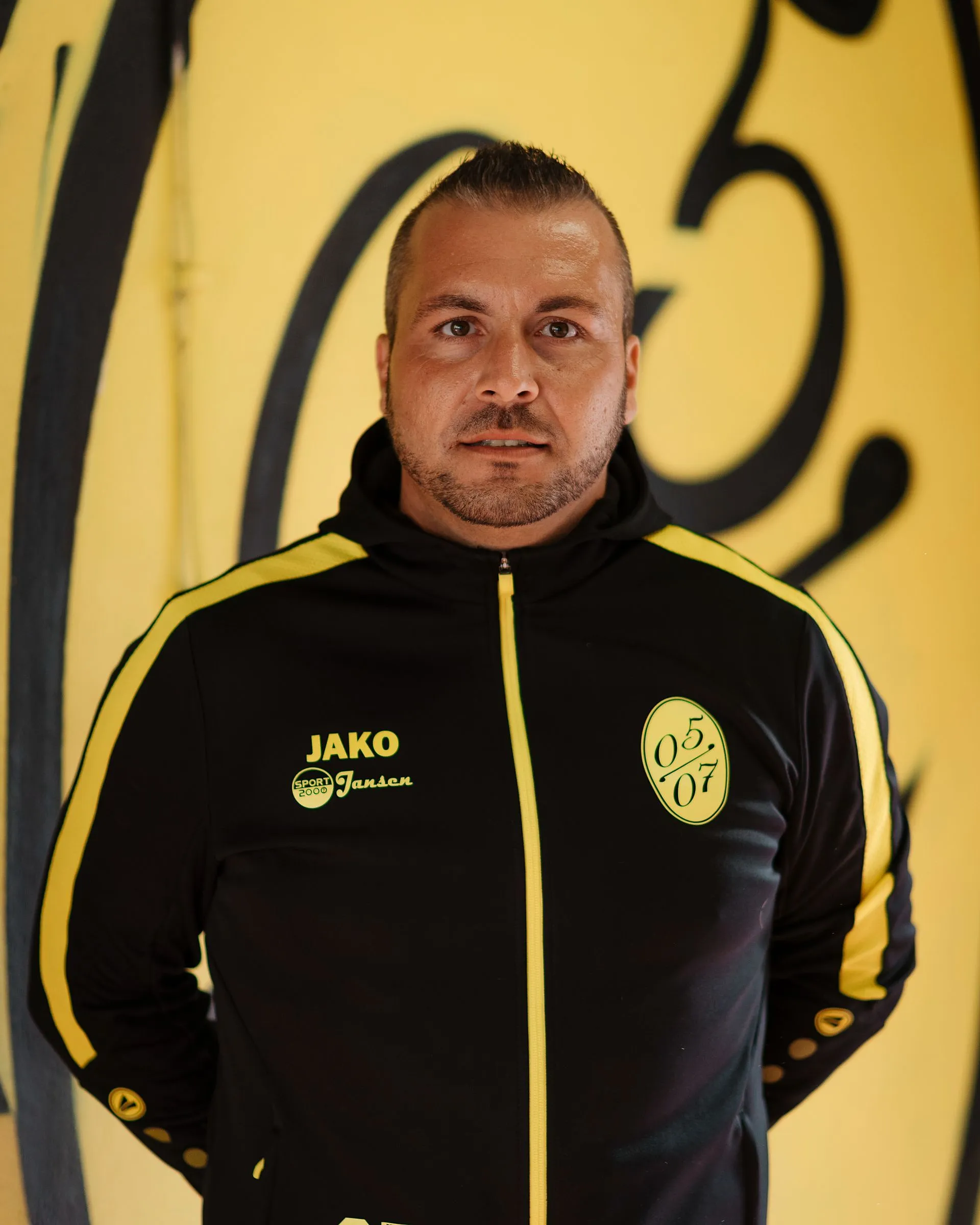 Ein Mann in einer schwarz-gelben Fußball-Herren-2-Sportjacke steht vor einer gelben Wand mit schwarzen Graffiti-Nummern und schaut mit neutralem Blick direkt in die Kamera.