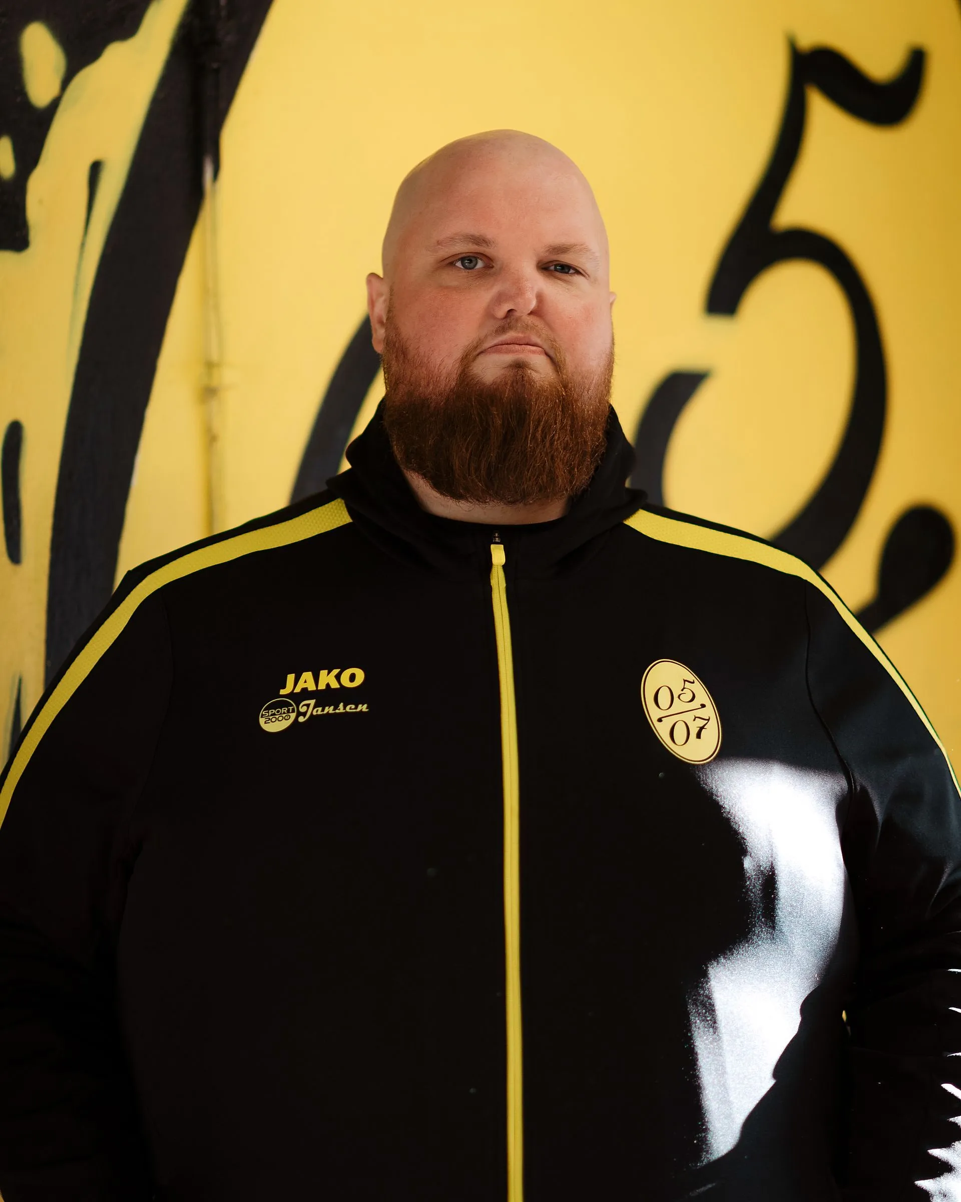 Ein bärtiger Mann in einer schwarz-gelben Fußball Herren 2 Sportjacke steht vor einer gelben Wand mit großen schwarzen Zahlen. Das Sonnenlicht hebt einen Teil seines Gesichts und seiner Jacke hervor.