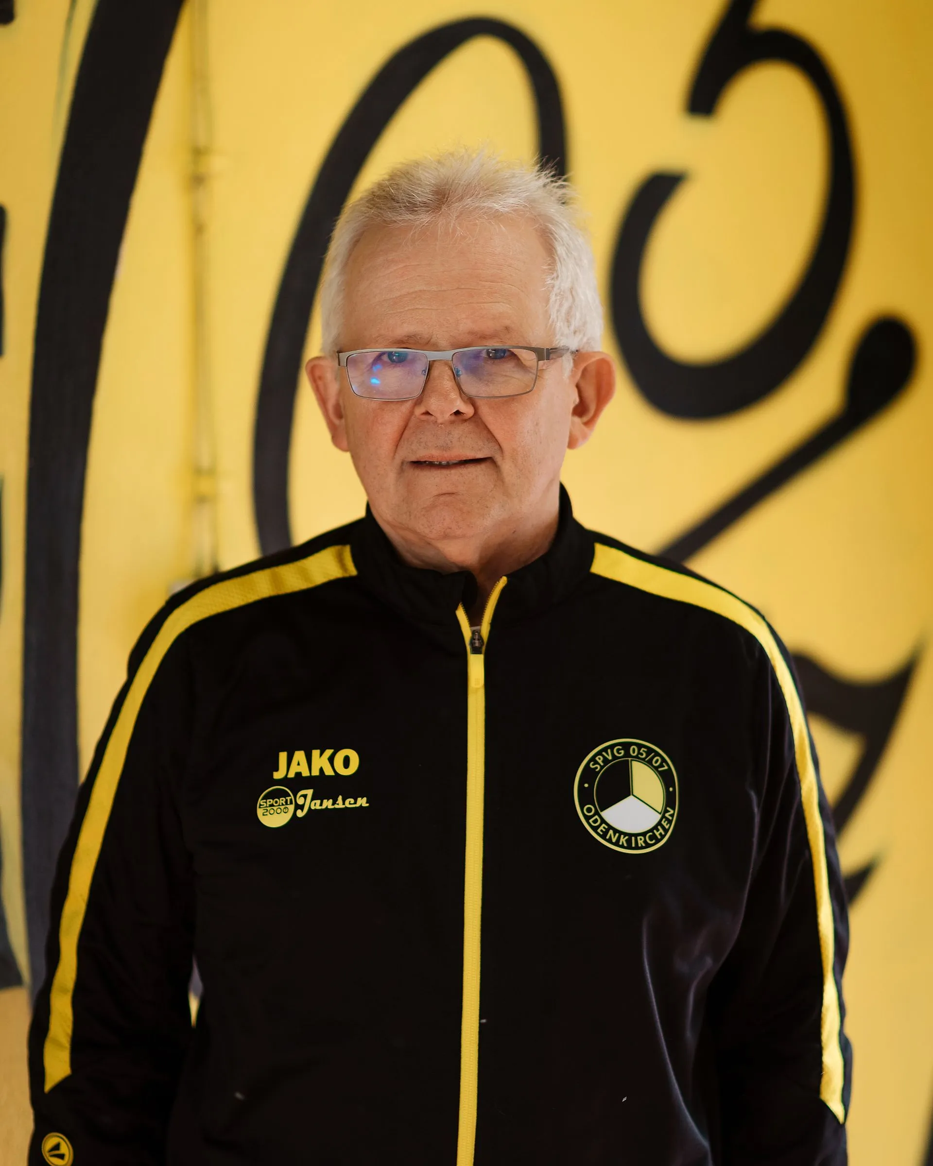 Ein älterer Mann mit kurzen grauen Haaren und Brille steht vor einer gelben Wand mit schwarzer Schrift. Er trägt eine schwarz-gelbe Sportjacke mit Logos, unter anderem Fußball Herren 2, sowie Jako und Jansen auf der rechten Seite.