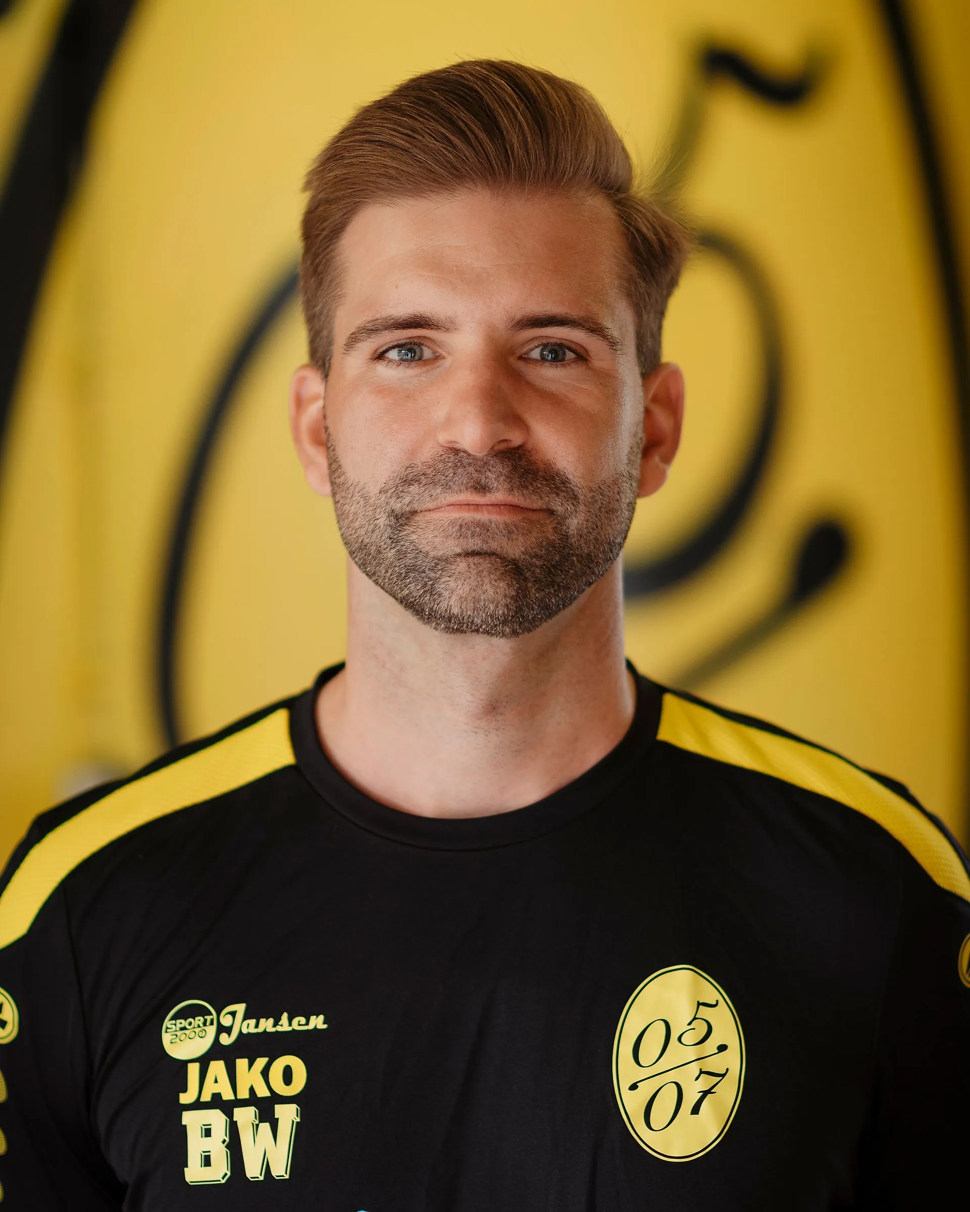 Ein Mann mit gepflegtem blondem Haar und gestutztem Bart trägt ein schwarz-gelbes Fußball Herren 3 Trikot mit den Logos "05 07", "JAKO" und "BW" und steht vor einem gelben Hintergrund mit einem großen verschwommenen Logo.