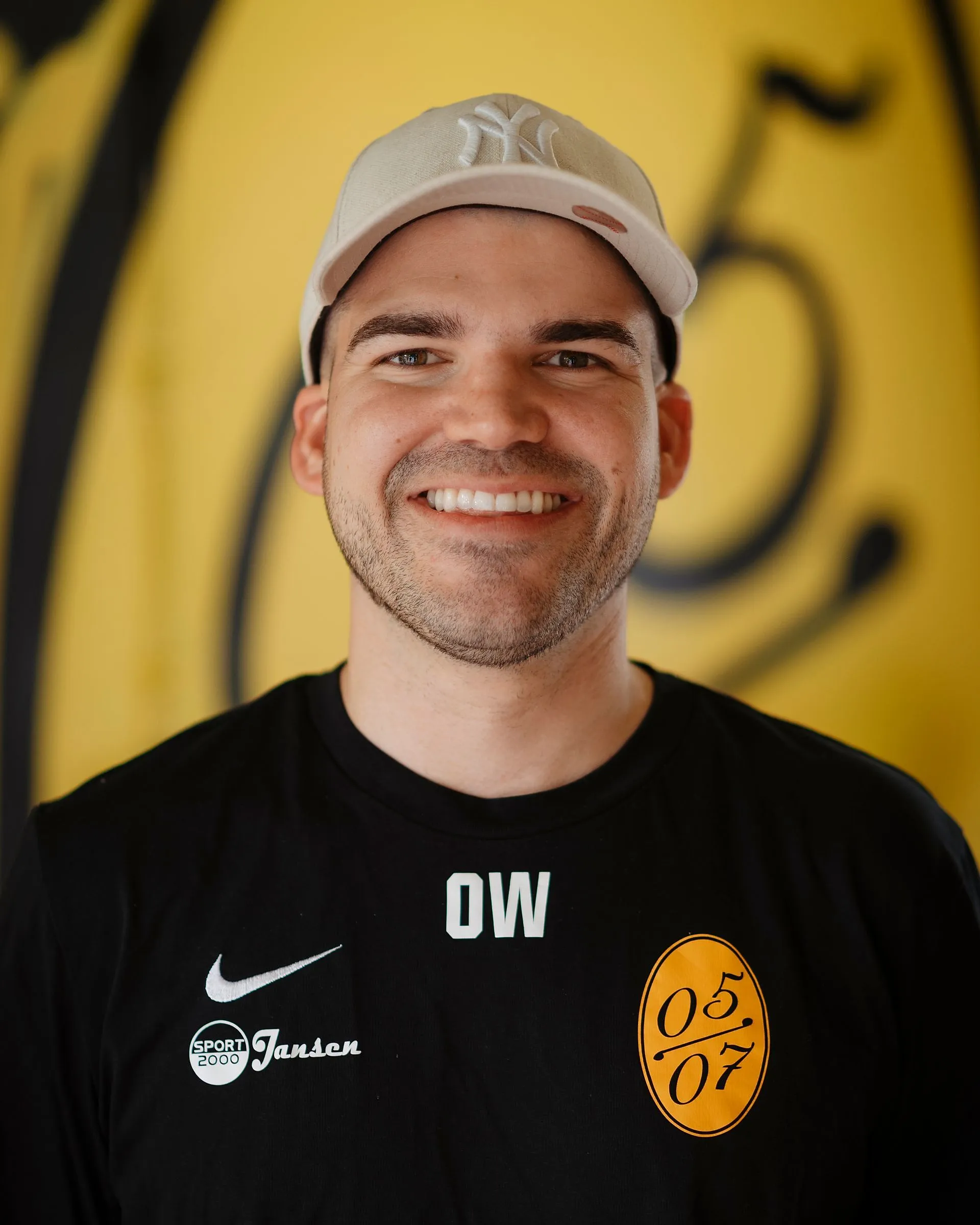 Ein Mann, der eine weiße Mütze und ein schwarzes Sporthemd mit weißem und gelbem Logo trägt, lächelt vor einem gelben Hintergrund mit schwarzen Markierungen, der Fußball Herren 3 darstellt.