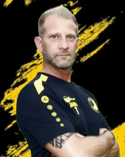 Ein Mann mit kurzen blonden Haaren und gestutztem Bart steht mit verschränkten Armen da, trägt ein schwarzes Sporthemd mit gelben Akzenten - eine Anspielung auf die Fußballjugend - und sein Armtattoo ist vor einem schwarzen Hintergrund mit gelben Pinselstrichen zu sehen.