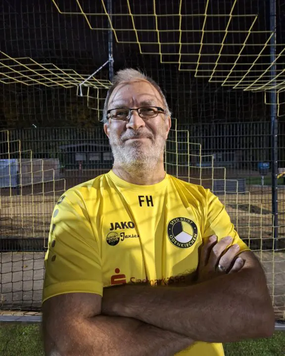 Ein Mann mit Brille und grauen Haaren steht selbstbewusst mit verschränkten Armen da und trägt ein gelbes FH Fußball Jugend Trikot. Er steht auf einem Fußballplatz, und im Hintergrund ist bei Nacht ein Netz zu sehen.
