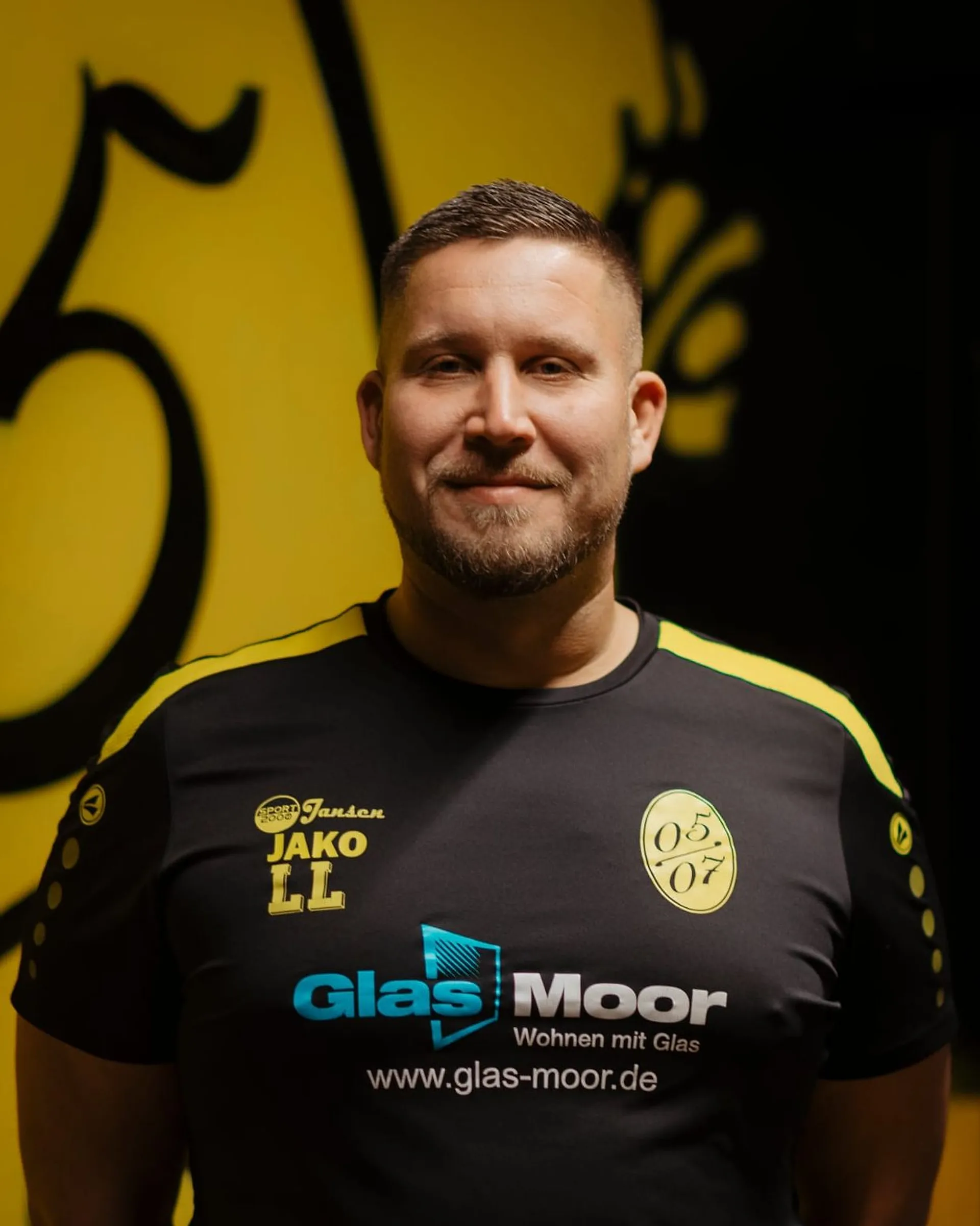 Ein Mann mit kurzen Haaren und Bart steht lächelnd da und trägt ein schwarz-gelbes Fußball-Jugend-Sporttrikot mit Sponsorenlogos und den Zahlen 05 07 auf der Brust. Im Hintergrund ist eine große gelbe Grafik zu sehen.