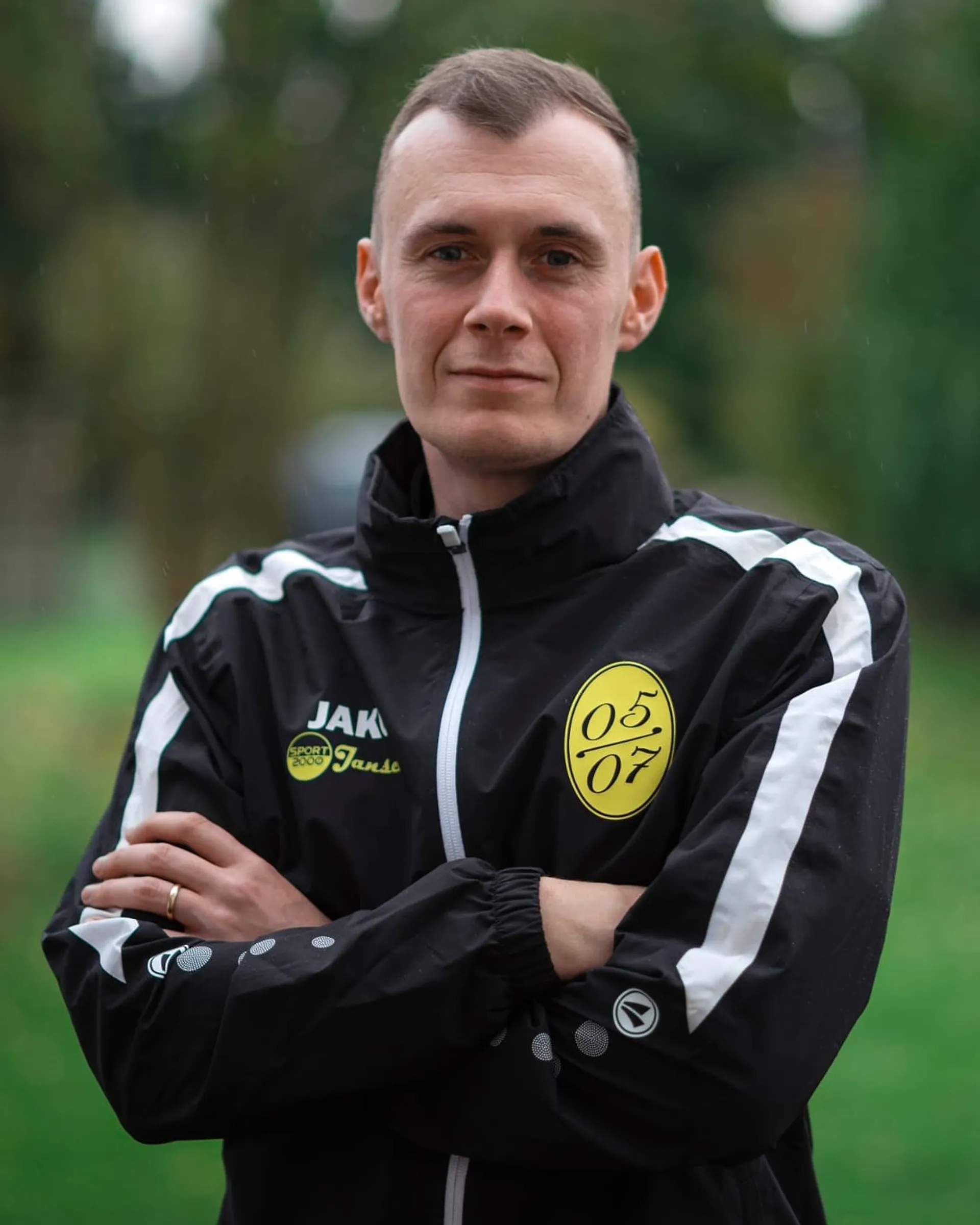 Ein Mann mit kurzen Haaren, der eine schwarz-weiße Fußball-Jugend-Sportjacke trägt, steht mit verschränkten Armen im Freien und schaut in die Kamera. Auf der Jacke sind gelbe kreisförmige Logos und der Name Janis zu sehen. Das Grün im Hintergrund ist unscharf.