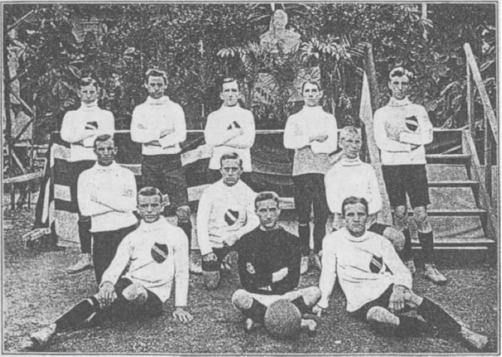 Ein altes Schwarz-Weiß-Foto für die Vereinschronik zeigt elf Männer in Fußballtrikots, die im Freien posieren - einige stehend, andere kniend oder sitzend, mit einem Fußball im Vordergrund und Pflanzen und Treppen im Hintergrund.