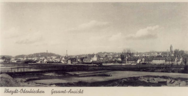 Ein altes Schwarz-Weiß-Panoramafoto von Rheydt-Odenkirchen, das in einer Vereinschronik abgebildet ist, zeigt eine Stadt mit verstreuten Gebäuden, Kirchtürmen, Schornsteinen und offenem Land im Vordergrund unter einem bewölkten Himmel.