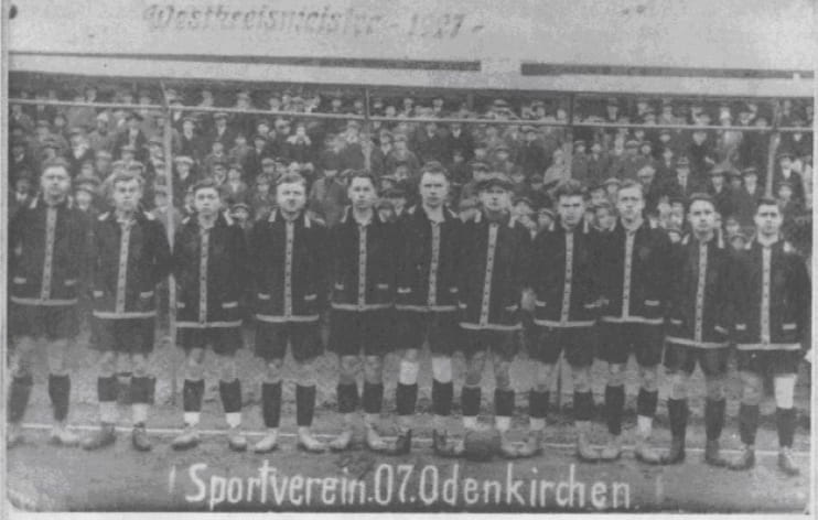 Ein altes Schwarz-Weiß-Foto aus der Vereinschronik zeigt eine Fußballmannschaft von elf Männern in dunklen Trikots mit hellen Verzierungen, die in einer Reihe auf dem Spielfeld stehen. Hinter ihnen sitzen Zuschauer auf Tribünen. Der Text am unteren Rand lautet Sportverein 07 Odenkirchen.