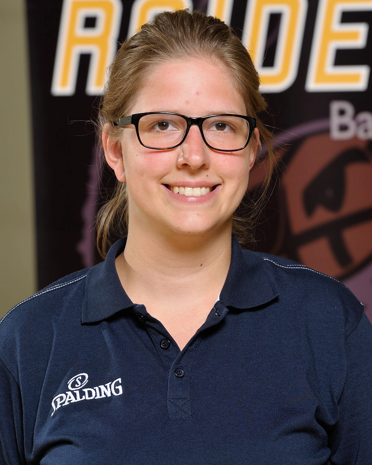 Eine Frau mit Brille und zurückgebundenem Haar lächelt in die Kamera. Sie trägt ein marineblaues Spalding-Poloshirt. Hinter ihr ist ein schwarz-gelbes Basketball-Raiders-Banner zu sehen, auf dem teilweise das Wort RAIDERS zu lesen ist.