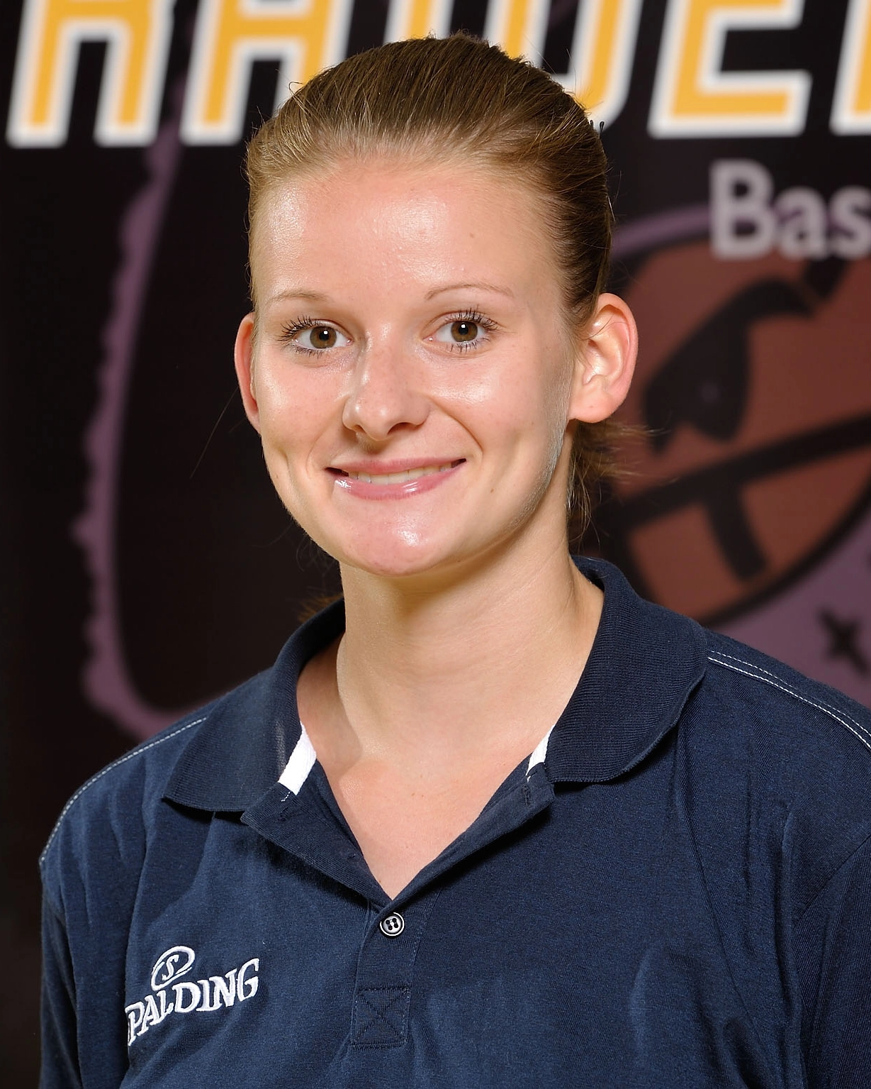 Eine junge Frau mit heller Haut und blondem, zurückgebundenem Haar, die ein marineblaues Spalding-Poloshirt trägt, lächelt in die Kamera. Der verschwommene Hintergrund enthält Text und ein Basketball Raiders-Logo, was auf eine Sport- oder Basketballumgebung hindeutet.