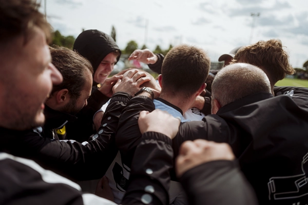Die Mannschaft von Fußball Odenkirchen 0507 steht im Freien eng beieinander, legt sich gegenseitig die Hände auf den Rücken und die Schultern und zeigt sich unter dem wolkenverhangenen Himmel mit den Bäumen im Hintergrund vereint und feierlich.