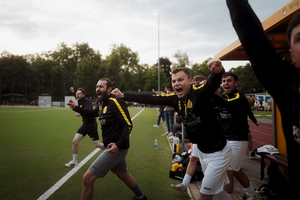Eine Gruppe von Spielern und Trainern von Fußball Odenkirchen 0507 in schwarz-gelben Trikots feiern begeistert an der Seitenlinie, heben die Fäuste und schreien vor Begeisterung während eines Spiels.