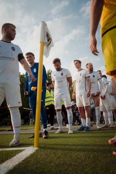 Eine Gruppe von Fußballspielern in weißen Trikots von Fußball Odenkirchen 0507 steht in einer Reihe in der Nähe einer Eckfahne auf dem Spielfeld, wobei ein Spieler in Gelb teilweise im Vordergrund zu sehen ist und ein blauer Himmel darüber liegt.