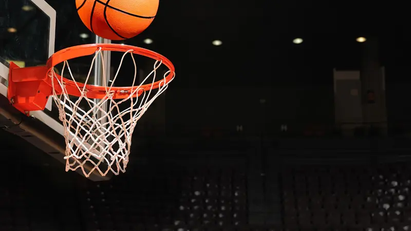 Ein Basketball Raiders-Ball fliegt durch einen Reifen mit weißem Netz in einer Halle, mit einem dunklen, verschwommenen Publikum im Hintergrund.