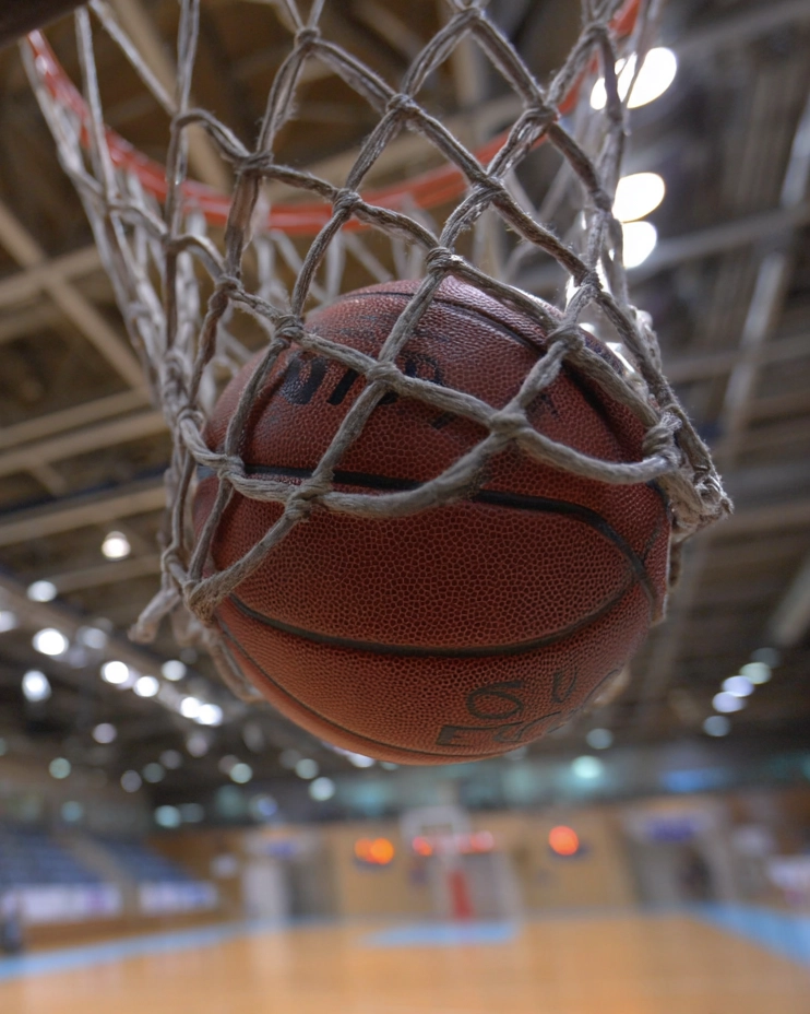 Ein Basketball, der sich aus einem niedrigen Winkel im Netz verfängt und kurz davor ist, den Korb in einer Halle der SpVg 0507 Odenkirchen zu treffen, mit unscharfer Beleuchtung und Sitzgelegenheiten im Hintergrund.