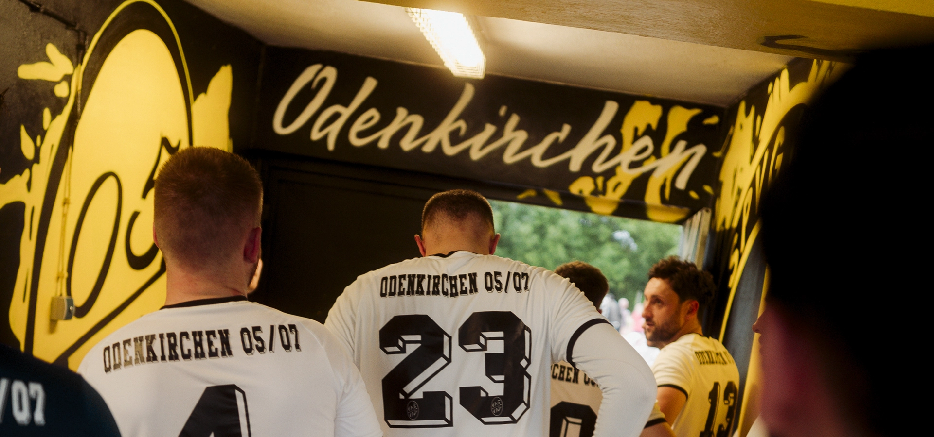 Mehrere Fußballspieler in weißen Trikots mit der Aufschrift "SpVg 0507 Odenkirchen" stehen in einem Tunnel unter einem passenden Schild und bereiten sich darauf vor, das Spielfeld zu betreten. Der Spieler in der Mitte trägt die Nummer 23.
