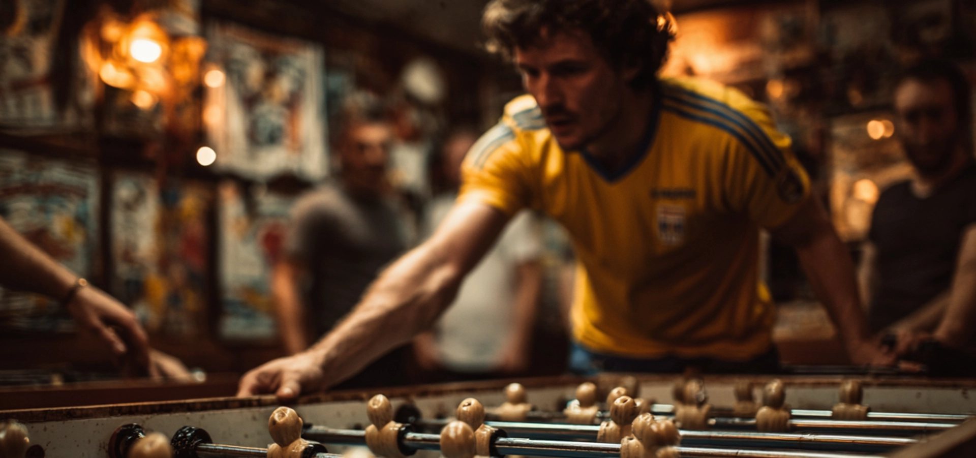 Ein Mann in einem gelben Trikot der SpVg 0507 Odenkirchen spielt in einem schwach beleuchteten Raum konzentriert Tischfußball, während andere Personen im Hintergrund verschwimmen und Plakate die Wände bedecken.