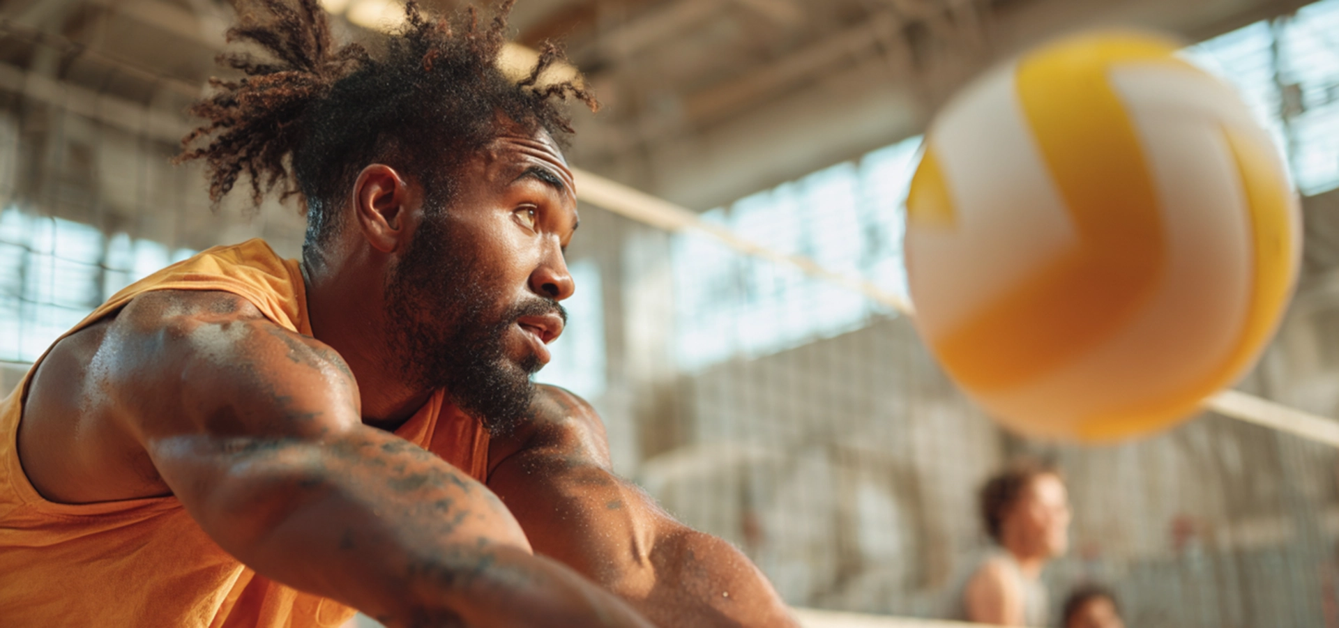 Ein konzentrierter männlicher Sportler der SpVg 0507 Odenkirchen in einem orangefarbenen Tank-Top bereitet sich darauf vor, einen gelb-weißen Volleyball in der Halle zu schlagen, mit unscharfen Spielern und einem Netz im Hintergrund.