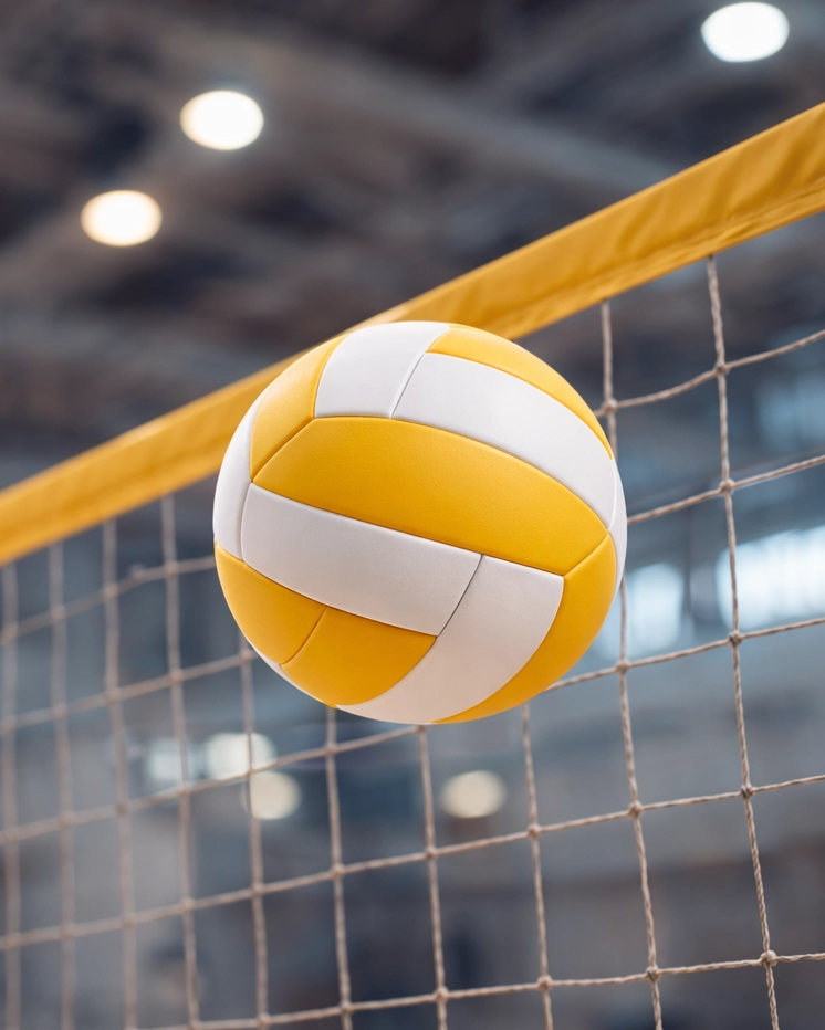 Ein gelb-weißer Volleyball fliegt während eines Spiels der SpVg 0507 Odenkirchen über ein Hallennetz, wobei der Hintergrund der Sporthalle unscharf und die helle Deckenbeleuchtung sichtbar ist.