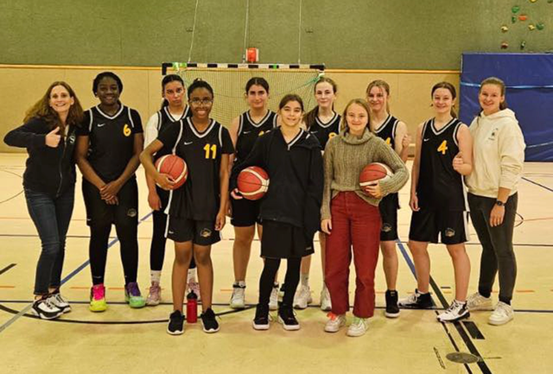 Eine Gruppe von zehn jungen Frauen, die meisten in schwarzen Basketball-Jugend-Uniformen, stehen zusammen auf einem Indoor-Basketballplatz. Drei halten Basketbälle in der Hand, und zwei in Freizeitkleidung stehen an den Enden, lächeln und posieren mit erhobenem Daumen.