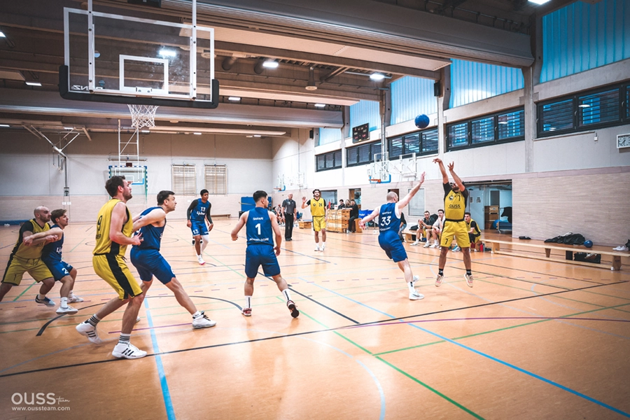 Eine Gruppe von Männern spielt ein Hallenbasketballspiel; ein Spieler der Basketball Raiders in einem gelben Trikot springt, um einen Wurf zu machen, während blau gekleidete Verteidiger versuchen, ihn zu blockieren. Andere Spieler schauen zu oder positionieren sich auf dem Spielfeld.