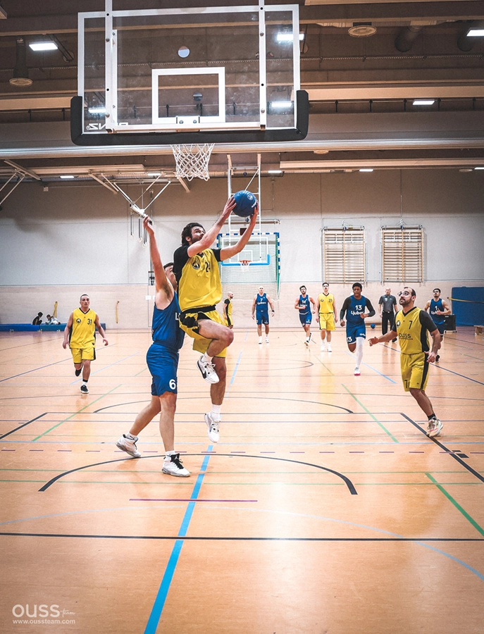 Ein Spieler der Basketball Raiders in einem gelben Trikot springt, um den Ball zu werfen, während ein Verteidiger in Blau versucht, ihn auf einem Hallenplatz zu blocken, während im Hintergrund Teamkollegen und ein Basketballkorb zu sehen sind.