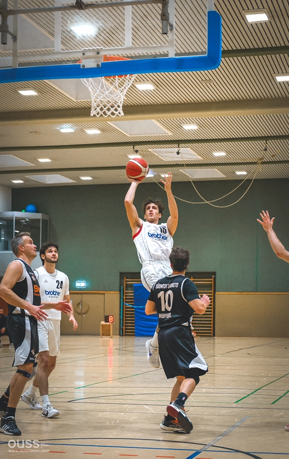 Ein Spieler der Basketball Raiders in einem weißen Trikot springt mit dem Ball in der Hand auf den Korb zu, während Gegner in schwarzen Trikots versuchen, ihn auf einem Indoor-Basketballplatz zu blockieren.