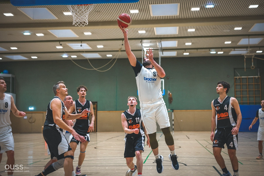 Ein Spieler der Basketball Raiders in einem weißen Trikot springt, um den Ball zu werfen, während vier Verteidiger in schwarzen Trikots und Mannschaftskameraden in einer Basketballhalle aufmerksam zusehen.