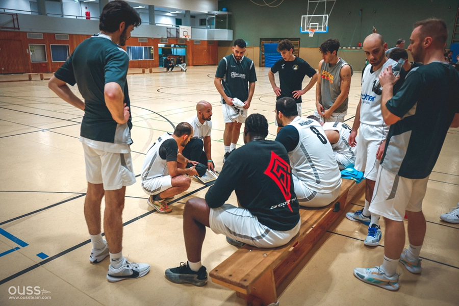 Ein Trainer der Basketball Raiders kniet auf dem Turnhallenboden und zeichnet auf einem Klemmbrett, während sich eine Gruppe männlicher Spieler in Trikots und Shorts um ihn herum versammelt und während einer Trainingseinheit aufmerksam zuhört.