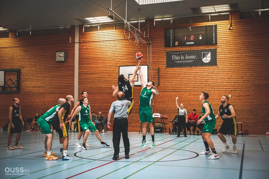 Die Spieler der Basketball Raiders springen während eines Spiels in einer Turnhalle um den Ball in der Mitte des Platzes. Der Schiedsrichter wirft den Ball zu, während andere Spieler und ein Trainer zuschauen. Im Hintergrund sind eine Anzeigetafel und ein "This is home"-Banner zu sehen.
