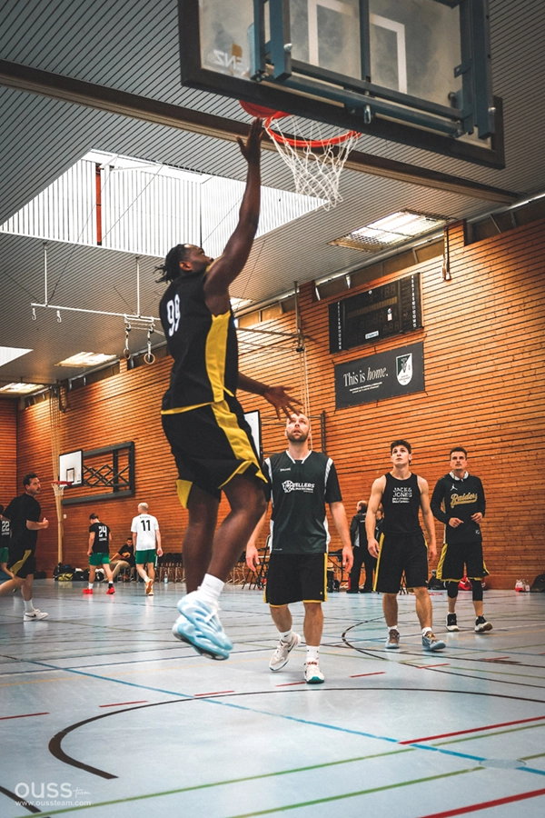 Ein Spieler der Basketball Raiders in einem schwarz-gelben Trikot springt zu einem Aufschlag, während vier andere auf einem Hallenplatz mit einer Ziegelwand und Bannern im Hintergrund zusehen.