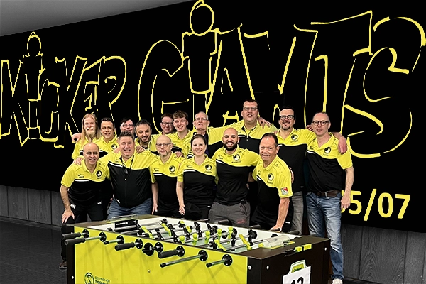 Eine Gruppe von Menschen in passenden gelb-schwarzen Hemden steht lächelnd hinter einem Tischfussballtisch, an dessen Wand in großen Buchstaben die Aufschrift Kicker Giants prangt.