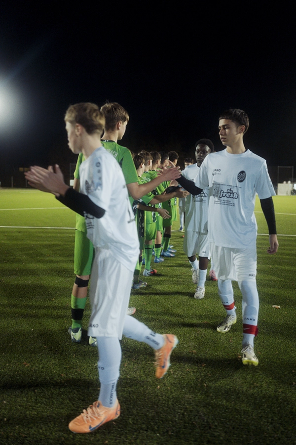 Zwei Jugendfußballmannschaften, darunter die SpVg 0507 Odenkirchen in grünen Trikots, stellen sich auf einem nächtlichen Spielfeld unter heller Stadionbeleuchtung auf und geben sich die Hand.