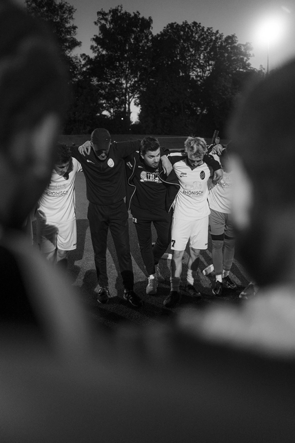 Ein Schwarz-Weiß-Foto der Fußballmannschaft der SpVg 0507 Odenkirchen, die eng aneinander gekuschelt auf dem Spielfeld steht, die Arme um die Schultern des anderen, mit Bäumen und Lichtern im Hintergrund.