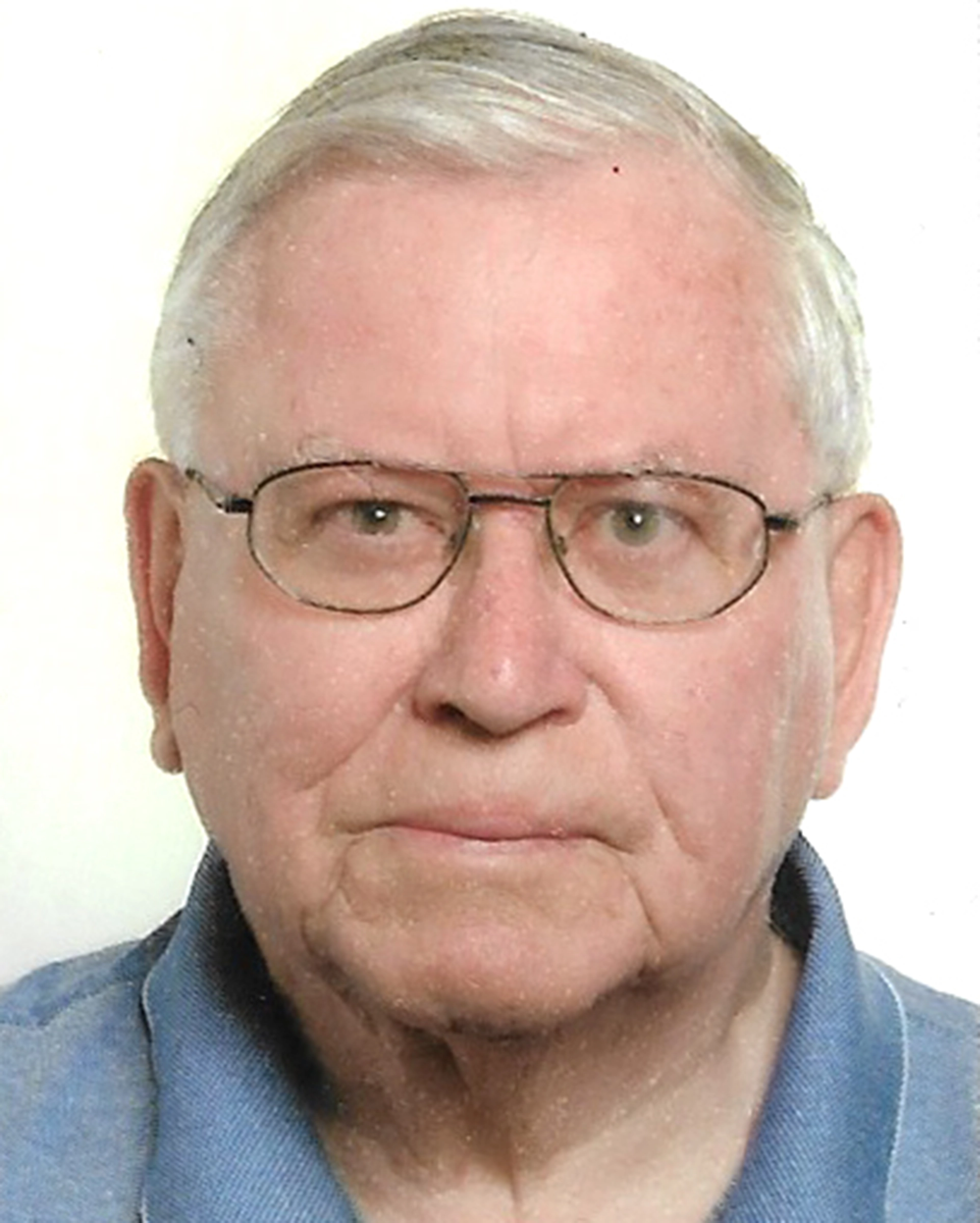 Ein älterer Mann mit kurzen grauen Haaren, der eine Brille und ein blaues Hemd mit Kragen trägt, blickt mit neutralem Gesichtsausdruck direkt in die Kamera - seine ruhige Haltung erinnert an ein Porträt des Präsidiums Kontakt, das vor einem schlichten weißen Hintergrund steht.